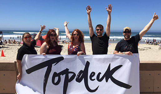 5 Topdeck employees holding up a flag of the Topdeck logo at the beach.