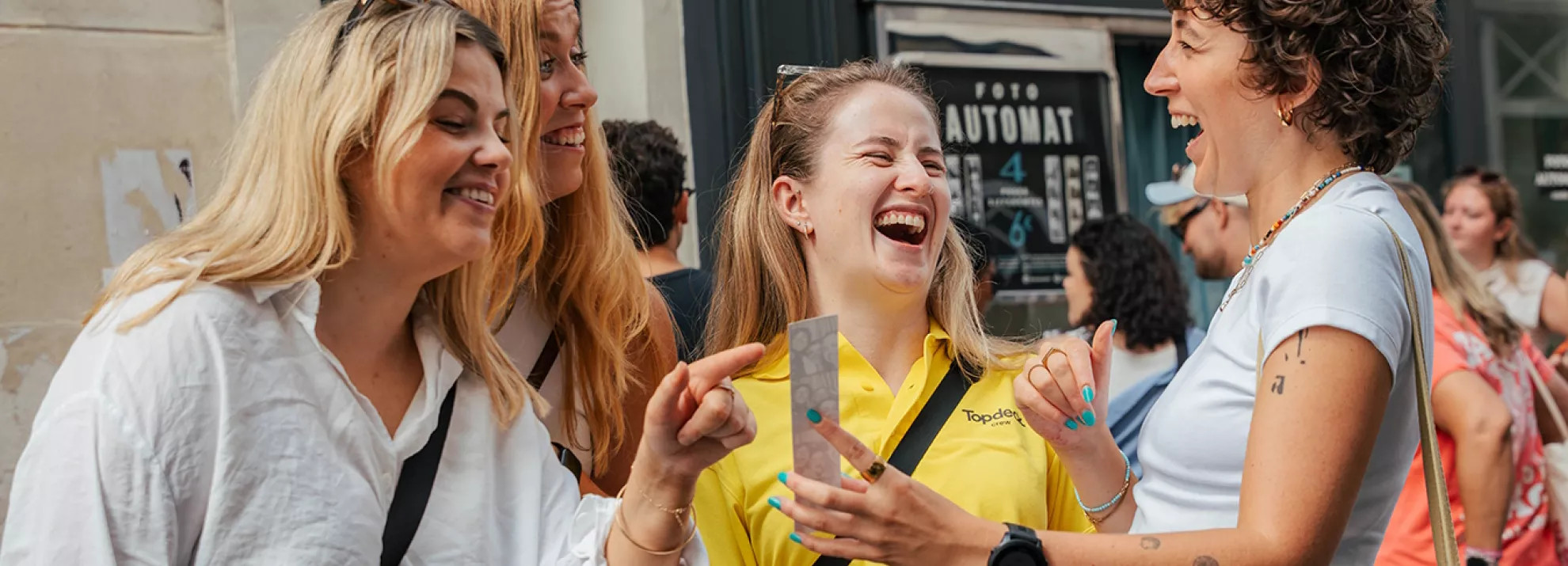 Four people laugh together while looking at a photo strip printed from a photobooth.