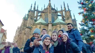 Group of people dressed in winter coats and smiling in front of St. Vitus Cathedral in Prague at Christmas time.