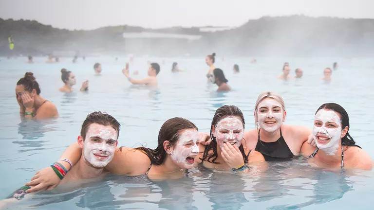 People with clay masks on their faces smile with their arms around each other in the Blue Lagoon.
