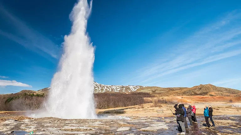 Geyser blowing hot water up into the air while a group of people stand to the right observing.