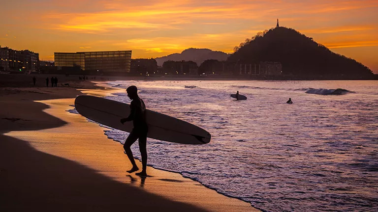 Surfer walking up the beach with their board as the sun sets in San Sebastian.