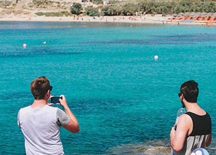 Topdeck travelers lounging in front of clear blue waters on a beach in Greece.