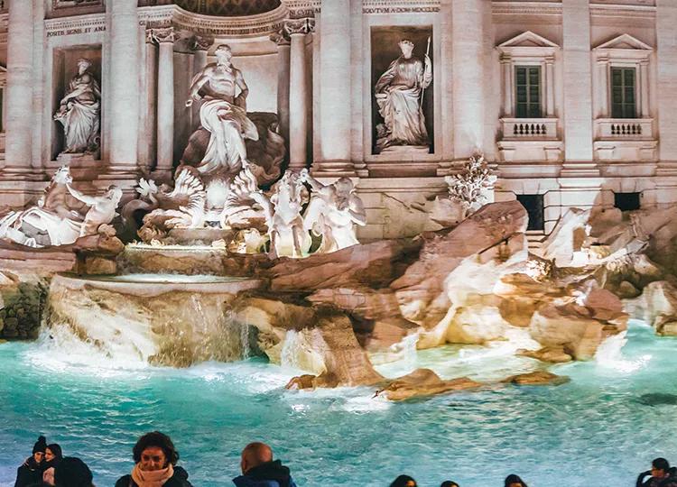 The famous Trevi Fountain of Rome lit up at night with a crowd of people surrounding it.