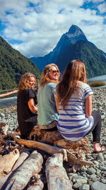Three women sit on drift-wood log while looking out at Milford Sound in New Zealand.